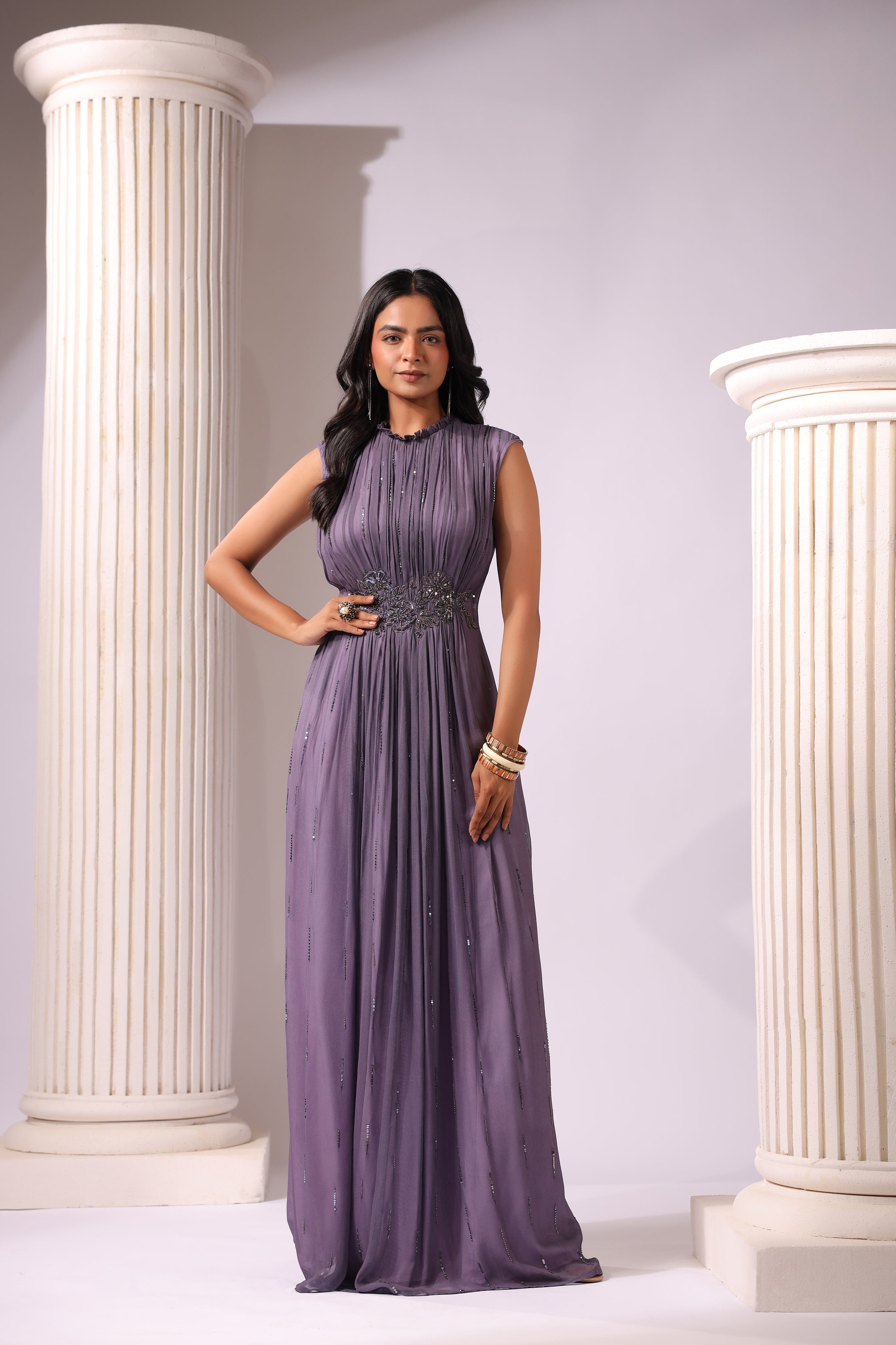 A woman standing in a classical columned setting wearing an elegant voilet gown with a side slit, accessorized with a belt and earrings.